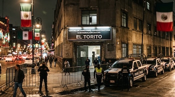 Fachada iluminada de un edificio con el letrero El Torito, patrullas, policías en la acera, vallas de seguridad y banderas mexicanas.