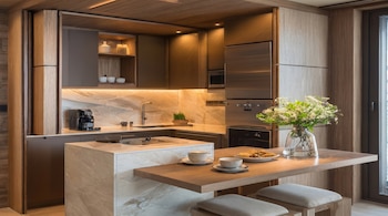 Cocina moderna con gabinetes de madera, encimeras de mármol y electrodomésticos integrados. Incluye una isla con estufa, barra con taburetes, flores y tazas.
