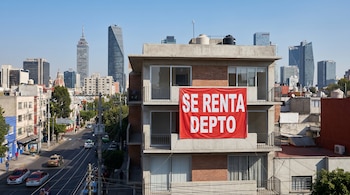 Edificio de departamentos en Ciudad de México con lona roja "SE RENTA DEPTO". De fondo, el skyline de la ciudad con la Torre Latinoamericana y otros edificios bajo cielo azul.