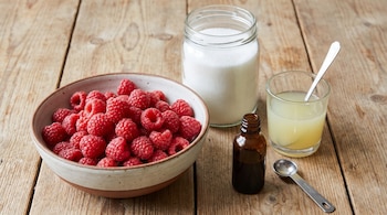 Un cuenco con frambuesas rojas, un frasco de vidrio con azúcar blanca, un vaso con jugo de limón y un frasco pequeño sobre madera.