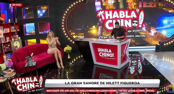 Milett Figueroa, con vestido brillante y tacones dorados, sentada en un sofá rojo en un set de televisión. A la derecha, un presentador en un escritorio