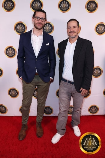Dos hombres en traje informal posan de pie sobre una alfombra roja frente a un fondo con logotipos de un evento de premios