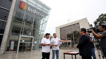 Dos hombres con papeles hablan frente al edificio de Indecopi con otro edificio de la Corte de Justicia de Lima de fondo. Un camarógrafo graba a un abogado.