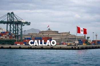 Un puerto con grúas, contenedores de carga, un cartel con la palabra CALLAO en mayúsculas, una bandera peruana y el Castillo Real Felipe al fondo.