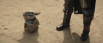 Grogu, una pequeña criatura verde con grandes orejas y ojos negros, de pie sobre arena. A su derecha, se ven las piernas blindadas de The Mandalorian