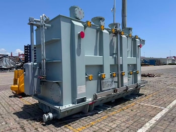 Vista exterior de un gran transformador eléctrico gris en un patio de adoquines, con un cielo azul claro y otros equipos industriales en el fondo