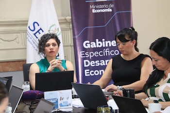 Tres mujeres sentadas en una mesa con laptops abiertas. Al fondo, una bandera con el logo 'SE' y un banner del Ministerio de Economía
