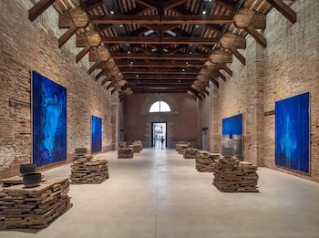 Interior de una galería con paredes de ladrillo y techo de madera. Se ven grandes cuadros azules en las paredes y esculturas de piedra apilada con cuencos negros en el suelo