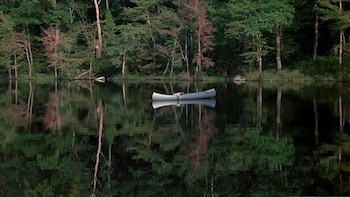 Una canoa blanca con una figura tumbada apenas visible flota en la superficie de un lago oscuro, que refleja un denso bosque de árboles verdes y rojizos