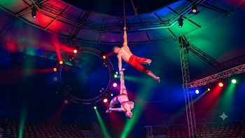 Dos artistas de circo realizan una acrobacia aérea colgados de una cuerda en el interior de una carpa, con luces de colores y asientos de público vacíos