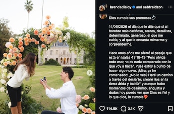 Una mujer y un hombre están arrodillados uno frente al otro en un jardín verde, bajo un arco de flores naranjas y blancas, frente a una mansión