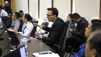 Un hombre con gafas y traje oscuro gesticula mientras habla en una sala de conferencias, rodeado de otras personas y laptops