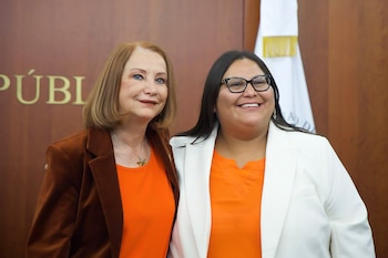 Primer plano de dos mujeres sonriendo. Una con cabello rojizo, chaqueta marrón; la otra con gafas, cabello oscuro y blazer blanco. Ambas visten tops naranja