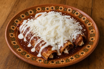 Plato de barro decorado con cuatro enchiladas en salsa roja, cubiertas con queso desmoronado, crema y tiras de cebolla, sobre madera.