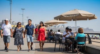 Vista panorámica de personas caminando y comiendo en una terraza junto a la playa en Lima bajo un cielo azul brillante; la costa y edificios se ven al fondo.