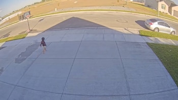 Niño descalzo corre en camino de entrada de concreto. En la calle hay un aro de baloncesto y un coche plateado. Hay pasto y casas a los lados