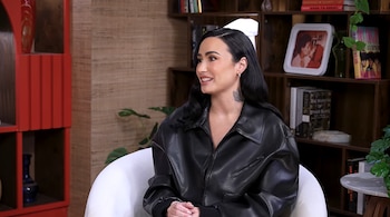 Demi Lovato sentada en una silla blanca con una chaqueta de cuero negra frente a una estantería de madera con libros y un portarretratos
