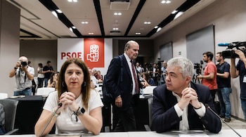 Dos personas en primer plano sentadas en una mesa con micrófonos. Detrás, el logo del PSOE y varias personas, incluyendo periodistas con cámaras.