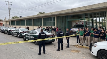 Patrullas, una ambulancia y policías detrás de una cinta de acordonamiento con estudiantes frente a un edificio con rejas verdes.