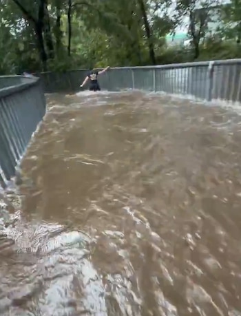 Un camino peatonal cubierto por agua turbia en movimiento, con barandillas metálicas en los laterales y una persona caminando al fondo