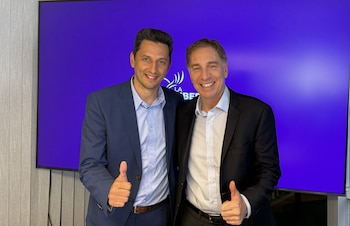 Dos hombres sonrientes, Diego Santilli y Adrián Núñez, con trajes, posan con el pulgar en alto frente a una pantalla morada con el logo de La Libertad Avanza