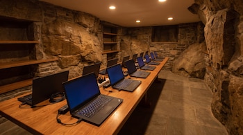 Diez computadoras portátiles encendidas y conectadas con cables descansan sobre una larga mesa de madera en una habitación con paredes de piedra.
