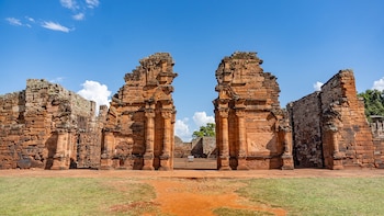 Vista frontal de las Ruinas de San Ignacio Miní, con dos grandes secciones de piedra rojiza y tallada flanqueando una apertura, sobre pasto verde y un cielo azul con nubes