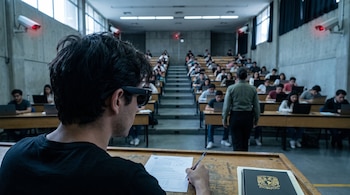 Vista trasera de un estudiante con lentes inteligentes en un pupitre con examen y bolígrafo. Aula universitaria con alumnos, supervisor y cámaras de vigilancia.