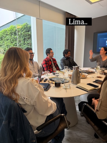 Personas sentadas alrededor de una mesa redonda con bebidas, notebooks y teléfonos, en una sala de reuniones con grandes ventanales y vegetación exterior