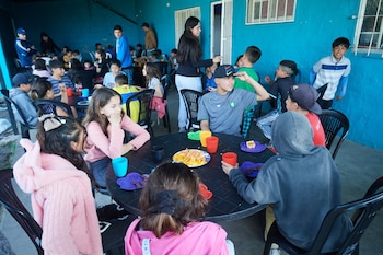 Varios niños y jóvenes sentados en mesas negras con tazas de colores, platos con fruta y postres. Las paredes son azules. Hay adultos de pie supervisando