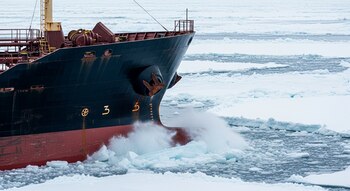 Primer plano de la proa de un buque petrolero oscuro con casco rojo, rompiendo hielo en aguas árticas. Trozos de hielo chocan contra la nave.