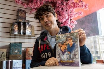 Bruno Pinasco, de cabello oscuro rizado y sudadera azul, sonríe a cámara mientras sostiene su libro "Tea Shop" en una librería con flores rosadas
