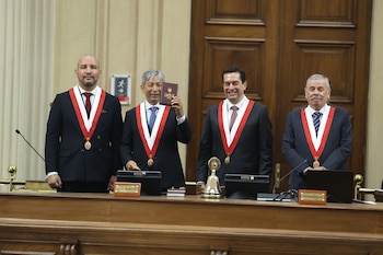 Cuatro hombres con traje oscuro y banda ceremonial roja y blanca están de pie detrás de un escritorio. Uno levanta un libro de la Constitución