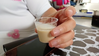 Mano de persona sosteniendo vaso de plástico transparente con líquido de dos capas, blanco y beige, sobre teléfono móvil negro y superficie estampada