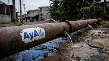 Una tubería de agua oxidada con el logo de AyA derrama agua por múltiples grietas, formando charcos en el suelo de un entorno urbano con vegetación tropical.