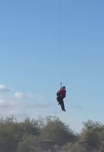 Un rescatista y una persona cuelgan de un cable suspendido en el aire sobre árboles y bajo un cielo despejado