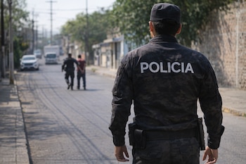 Vista trasera de un policía con uniforme camuflado oscuro y la palabra POLICIA en la espalda, observando una detención borrosa de una persona en una calle.