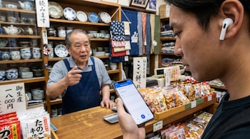 Primer plano de un cliente usando AirPods y un celular con Google Translate para interactuar con un tendero japonés en una tienda tradicional de Japón.
