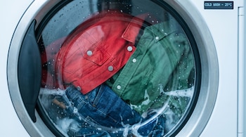 Lavadora de carga frontal con chaqueta de jean roja, camisa de algodón verde y pantalón de jean azul oscuro lavándose en agua fría a 25 grados Celsius.