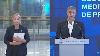 Un hombre de cabello gris en traje oscuro y camisa a cuadros habla desde un podio de Buenos Aires Ciudad. A la izquierda, una mujer rubia con blazer sostiene papeles