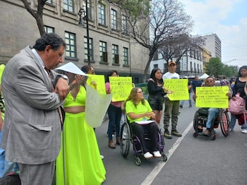 Un grupo de personas, incluyendo algunas en sillas de ruedas, manifiestan en una calle urbana. Sostienen pancartas con mensajes de protesta