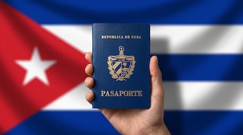 Una mano sostiene un pasaporte azul con letras y escudo dorados de Cuba ante la bandera cubana.
