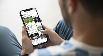 Hombre de espaldas con camiseta de Argentina. Sostiene un smartphone viendo su pantalla con publicaciones de redes sociales sobre un partido de fútbol.