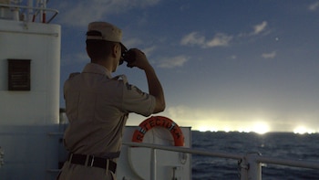 Vista trasera de un oficial con uniforme color caqui y gorra, mirando con binoculares hacia el océano con el horizonte iluminado por el sol