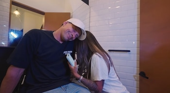 Hombre con gorra blanca, camiseta negra y pantalón vaquero, recostado sobre mujer con pelo largo y camiseta clara que sostiene un teléfono celular, con azulejos y puerta