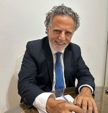 Hombre sonriente de mediana edad, con cabello rizado gris y barba, vestido con traje oscuro y corbata azul, sentado en un escritorio escribiendo en papel