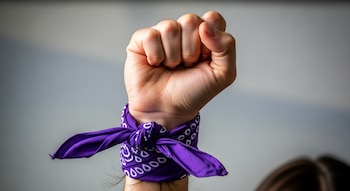 Primer plano de un puño femenino alzado con un pañuelo morado de patrón blanco anudado alrededor de la muñeca. El fondo es liso y gris claro.