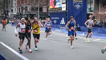 Tres corredores en una calle urbana, uno al centro es apoyado por otros dos. Al fondo se ven edificios, más corredores y pancartas del Maratón de Boston