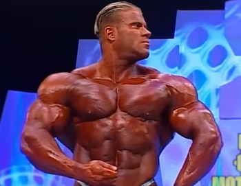 Hombre musculoso de torso desnudo posa en un escenario con fondo azul y morado, con luces y cabello rubio engominado hacia atrás