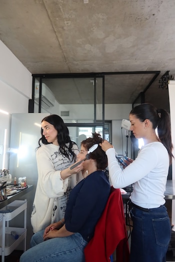 Tres mujeres en una habitación; una permanece sentada mientras otra le aplica maquillaje en el rostro y una tercera le peina el cabello
